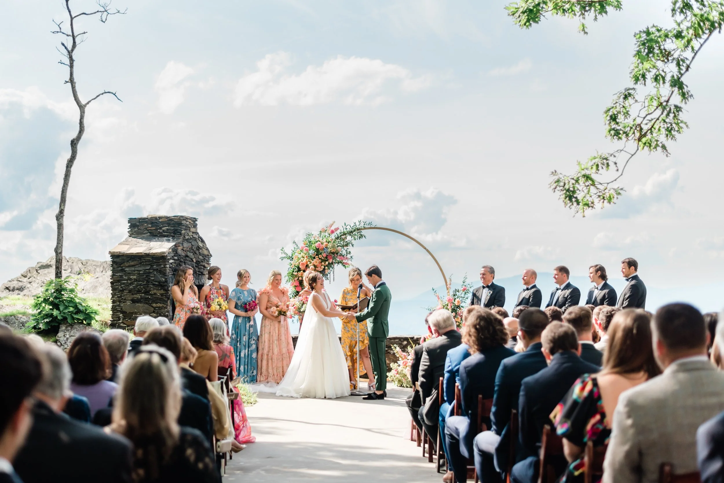 Wide view of an outdoor wedding ceremony with guests seated on both sides and the couple at the altar under a floral arch.