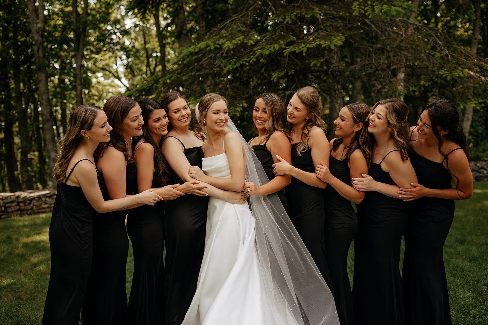 Bride smiling with bridesmaids wearing black dresses during a wooded outdoor wedding portrait.