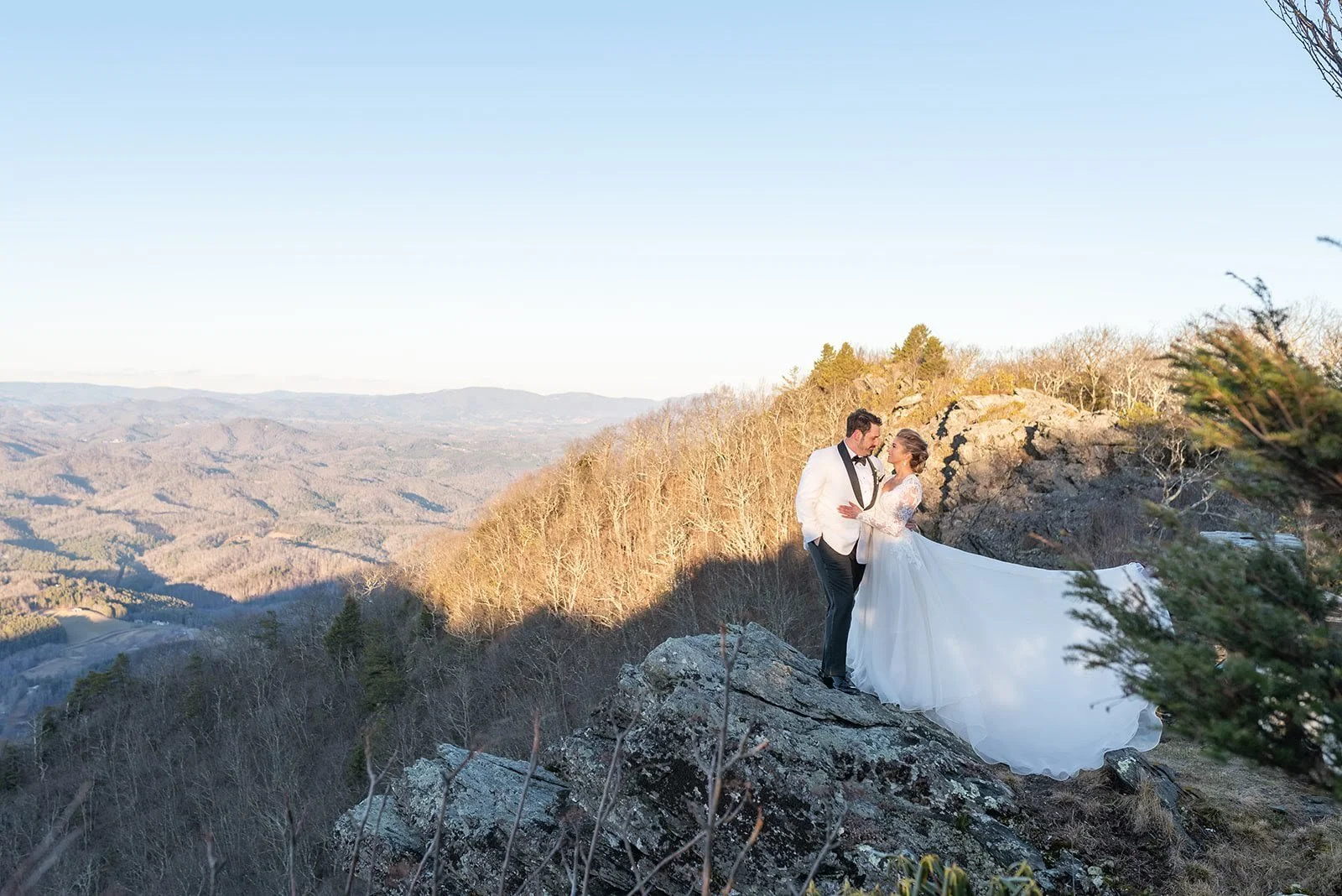 Wide mountaintop portrait of the bride and groom standing on a rocky cliff with sweeping mountain views.