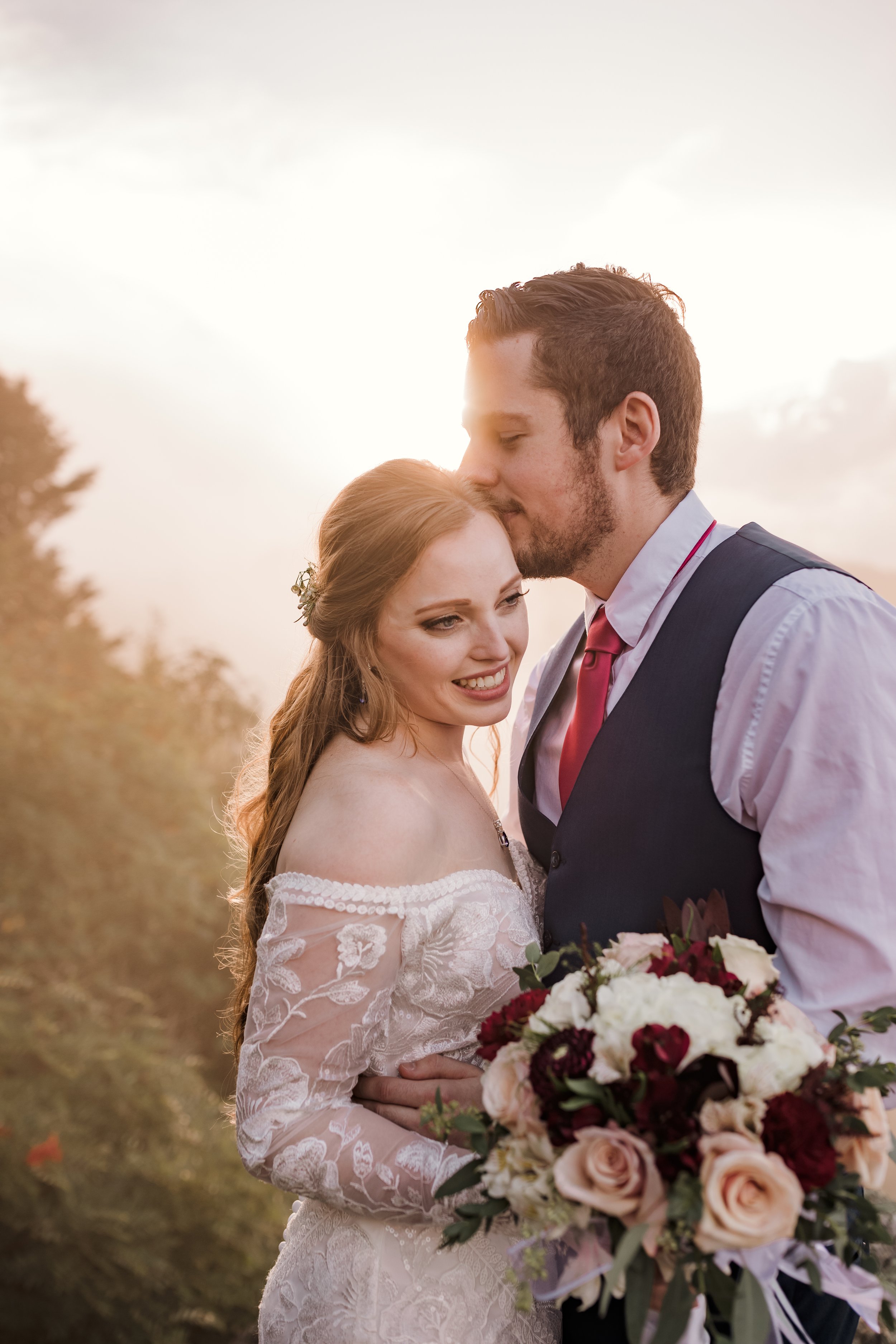 A groom in a vest and red tie kisses the forehead of his bride.