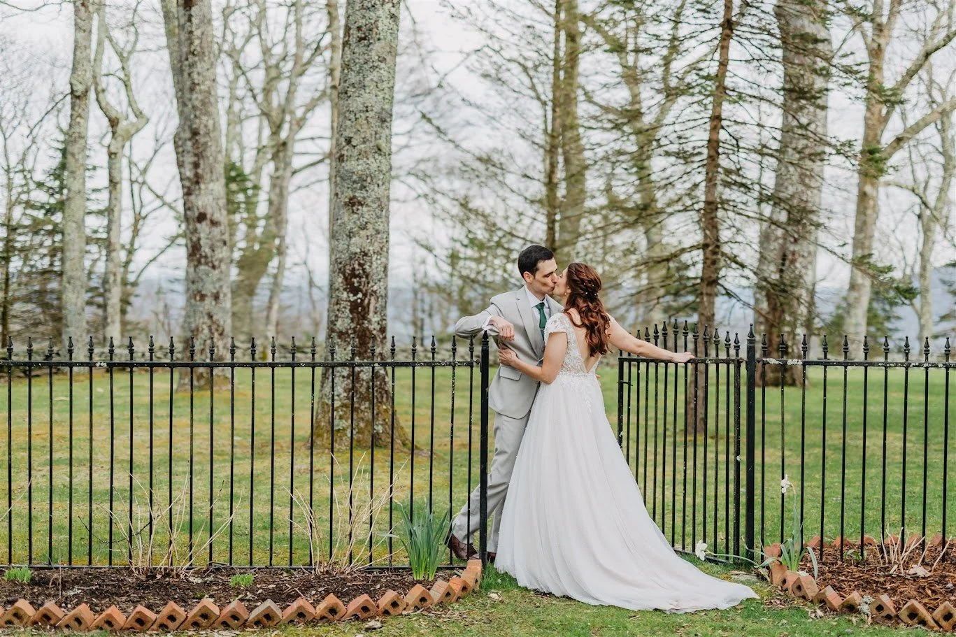 Bride and groom kissing by a black iron fence with tall trees in the backgroundBride and groom kissing by a black iron fence with tall trees in the background