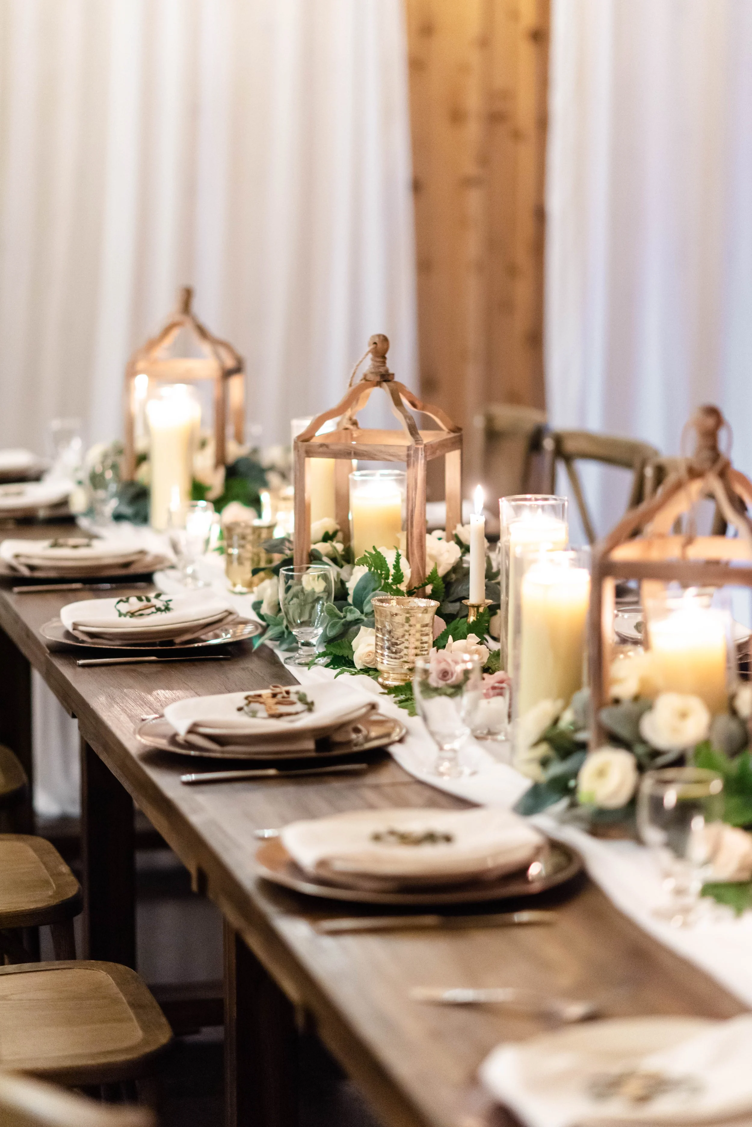 Romantic wedding reception table with candles, lanterns, greenery, white roses, and neutral place settings.