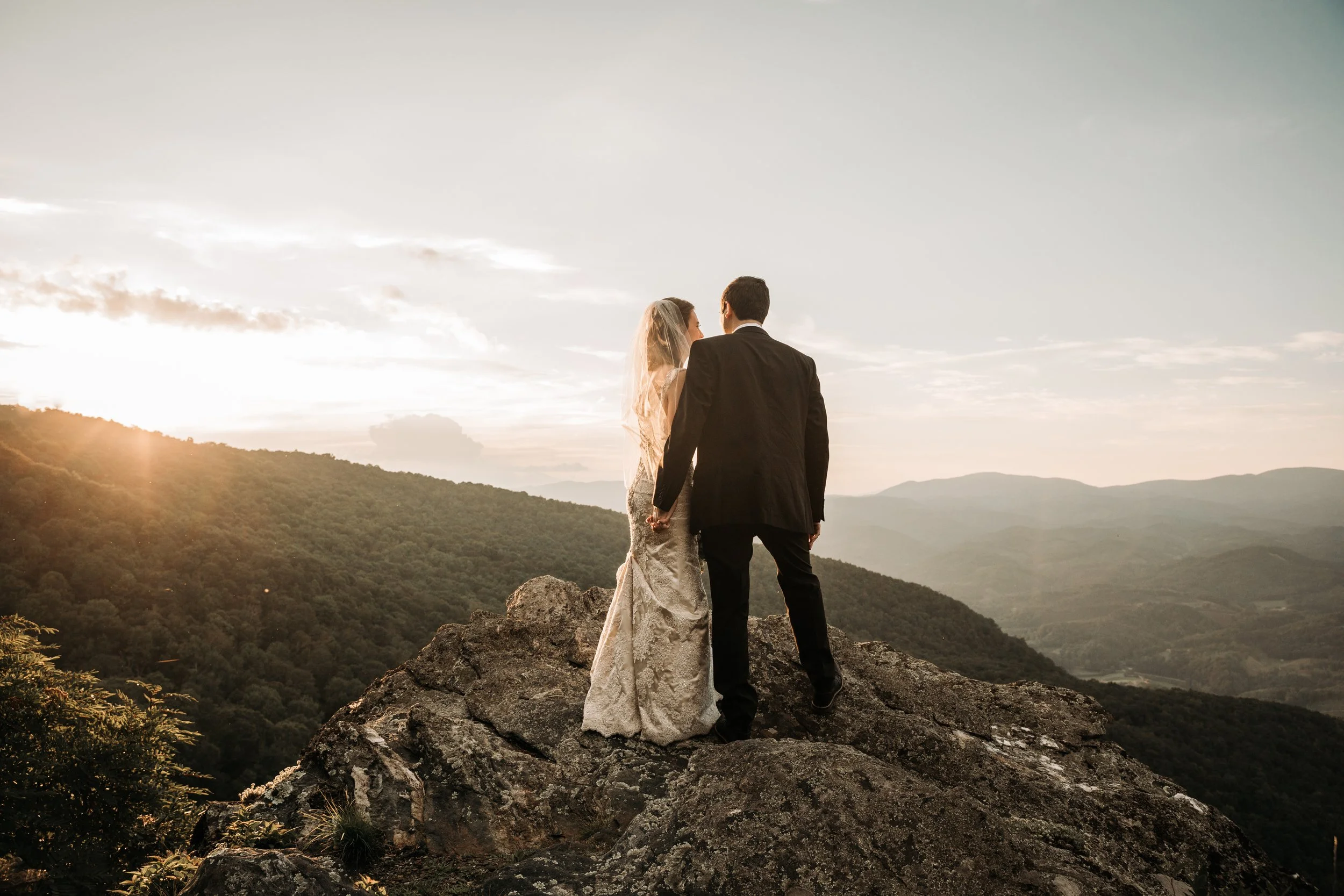 Newlyweds standing together on a mountain cliff at sunset with sweeping valley views.