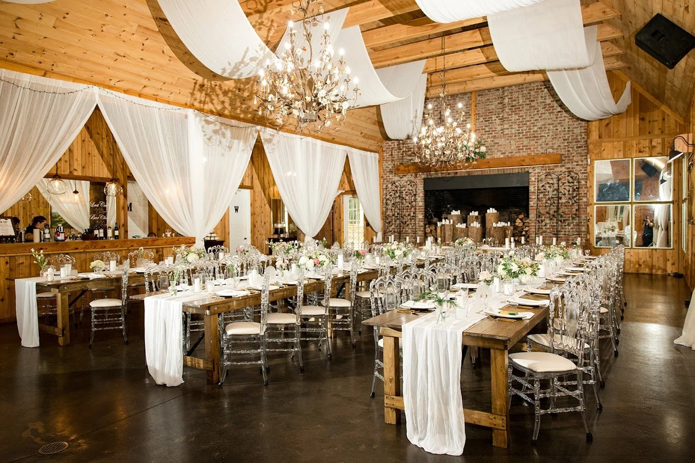 Wedding reception room with long wooden tables, clear chairs, white draping, and chandeliers