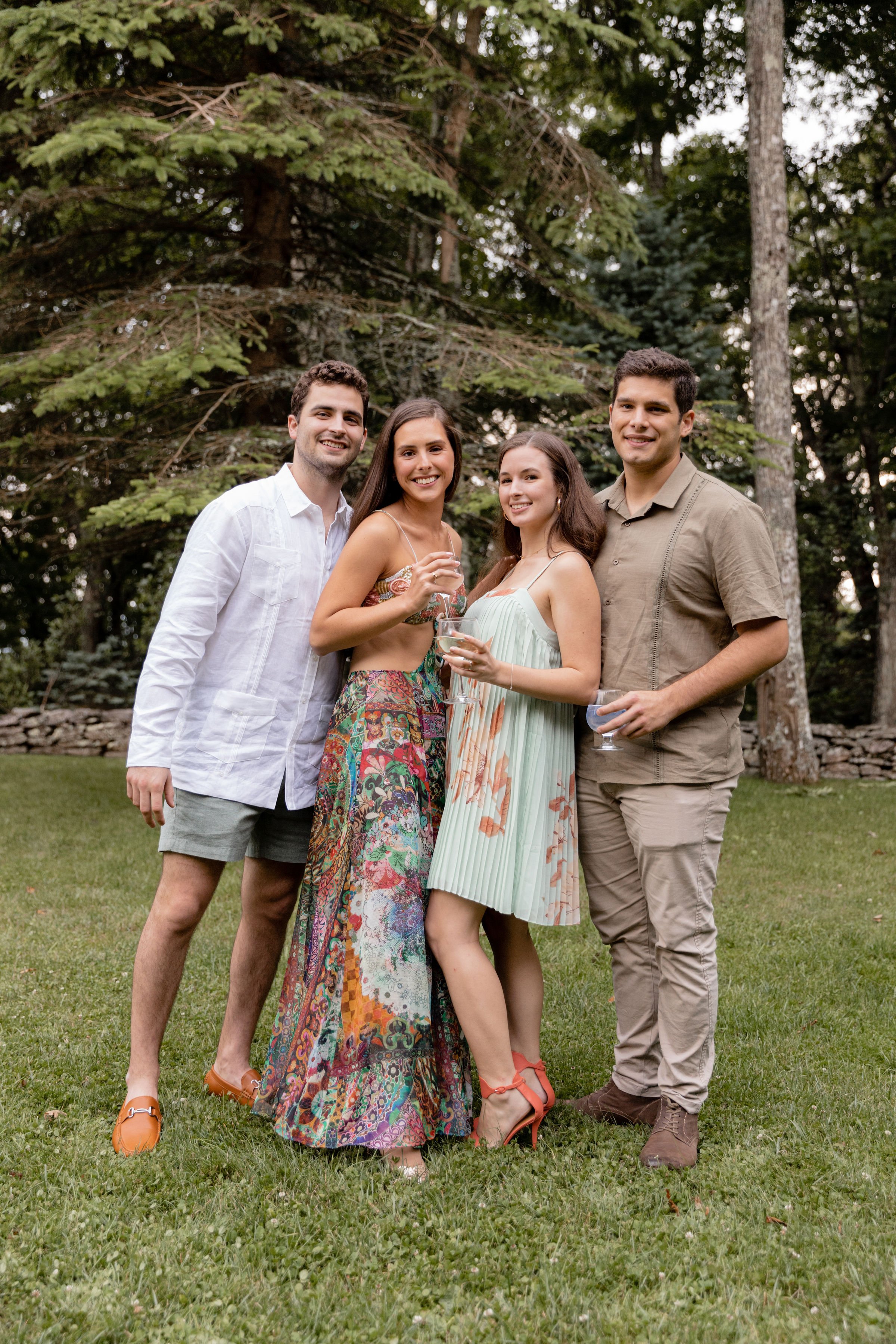 Friends posing with drinks on a lawn during a relaxed wedding weekend celebration surrounded by trees.