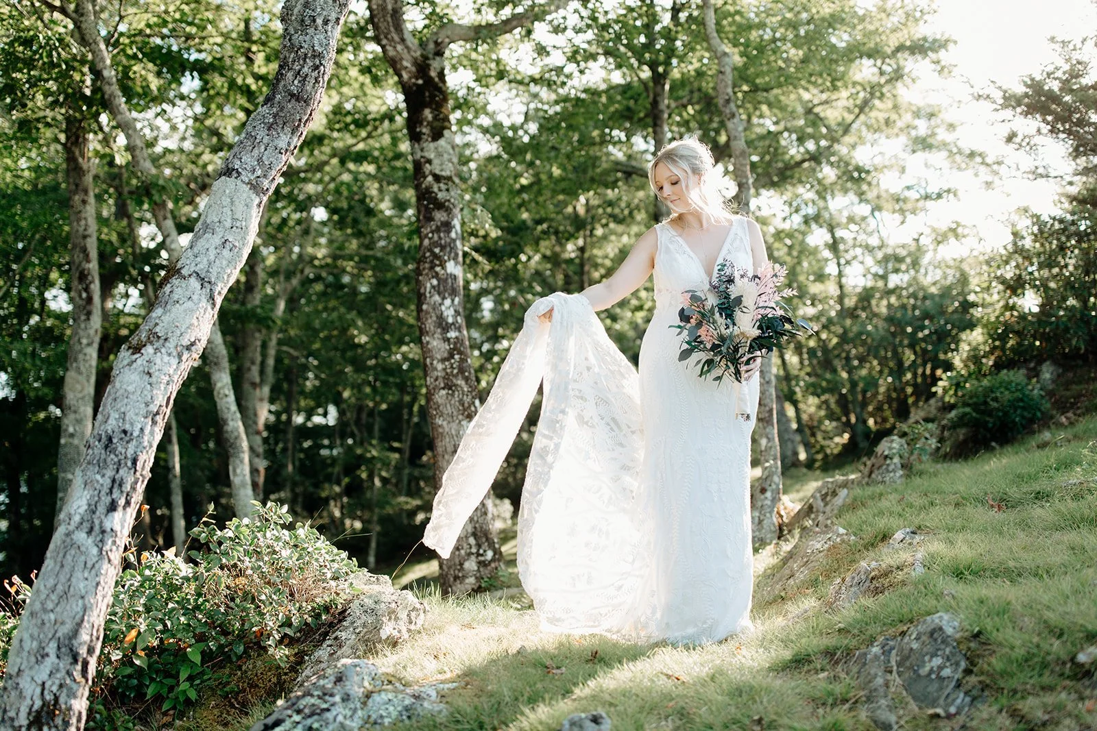 Bride standing in sunlit woods wearing a lace wedding dress and holding a moody floral bouquet.