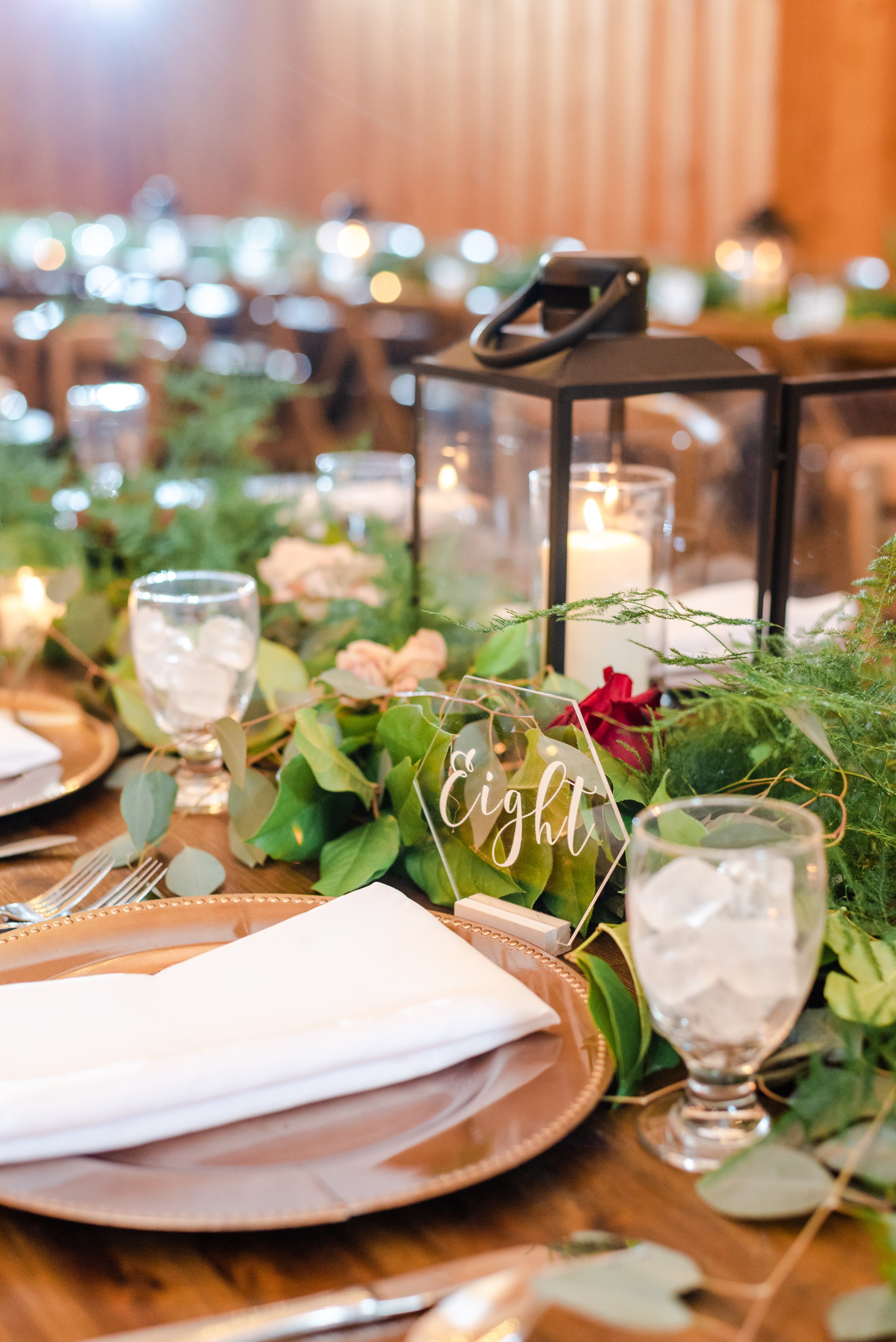 Wedding reception table setting with greenery runner, black lantern candles, and a table number sign.
