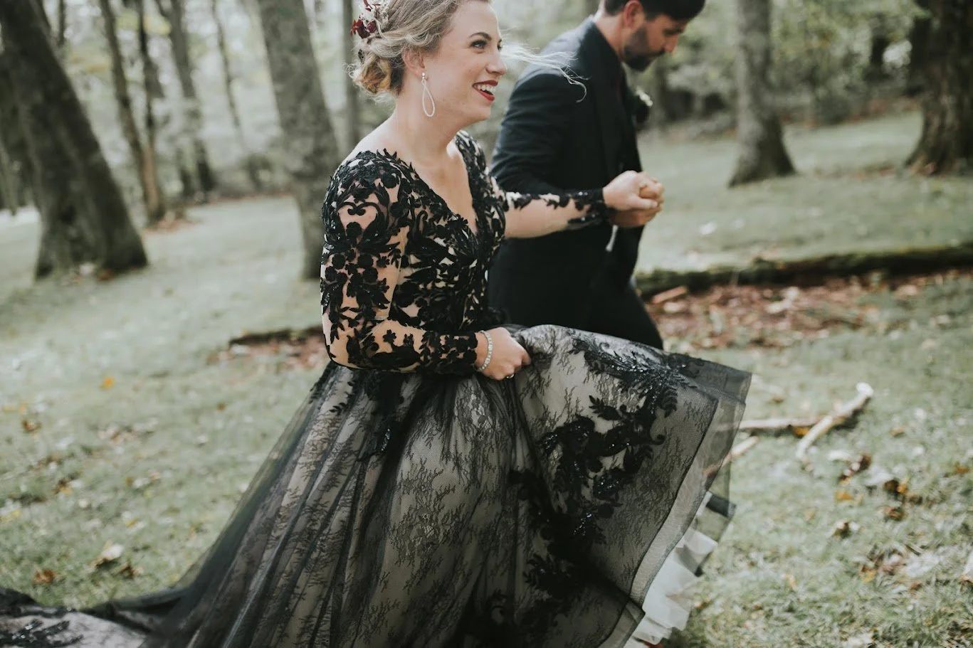 Bride in a black lace wedding gown running and smiling in a wooded outdoor setting.