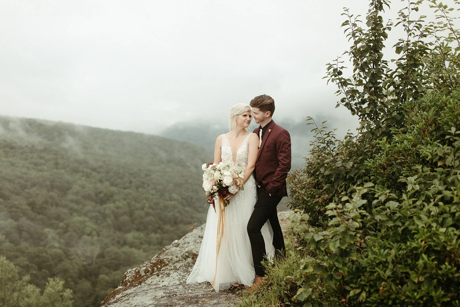 Bride and groom standing on a rocky ledge with a misty mountain forest view behind them