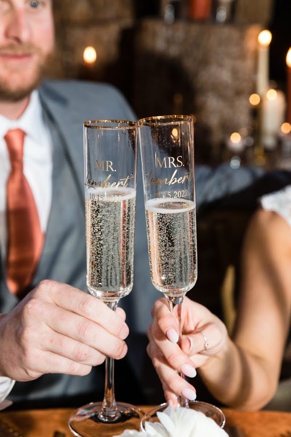 Wedding toast close-up with two champagne flutes clinking, “Mr” and “Mrs” text on the glasses.