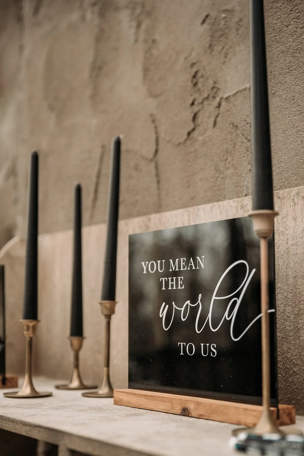 Black wedding sign reading you mean the world to us displayed with taper candles on a mantel.