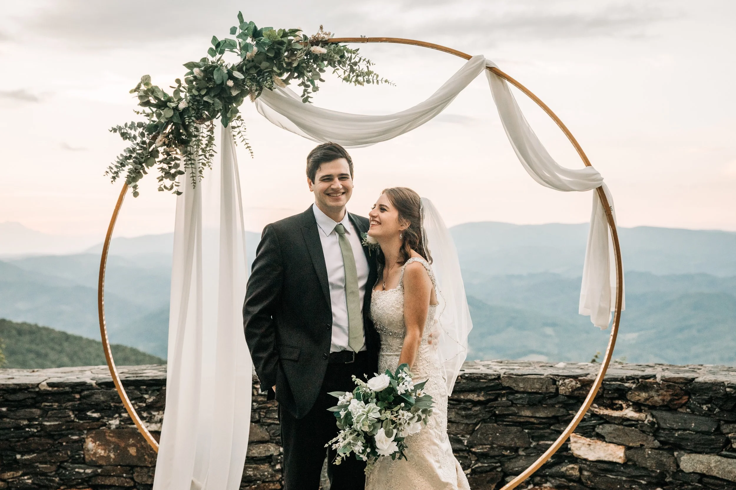Bride and groom standing beneath a circular wedding arch with soft mountain views in the background.