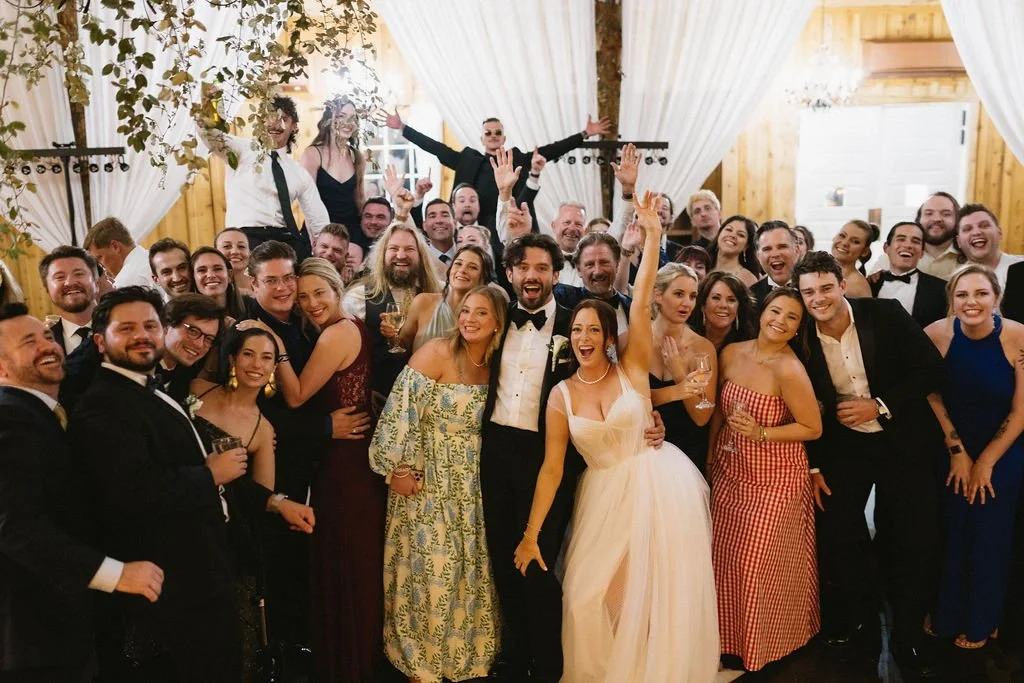 Bride and groom celebrating with a large group of wedding guests on the reception dance floor beneath white draping.