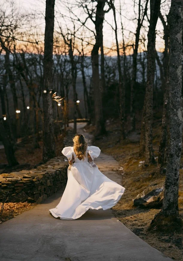 Bride walking down a wooded path at sunset with her dress flowing behind.