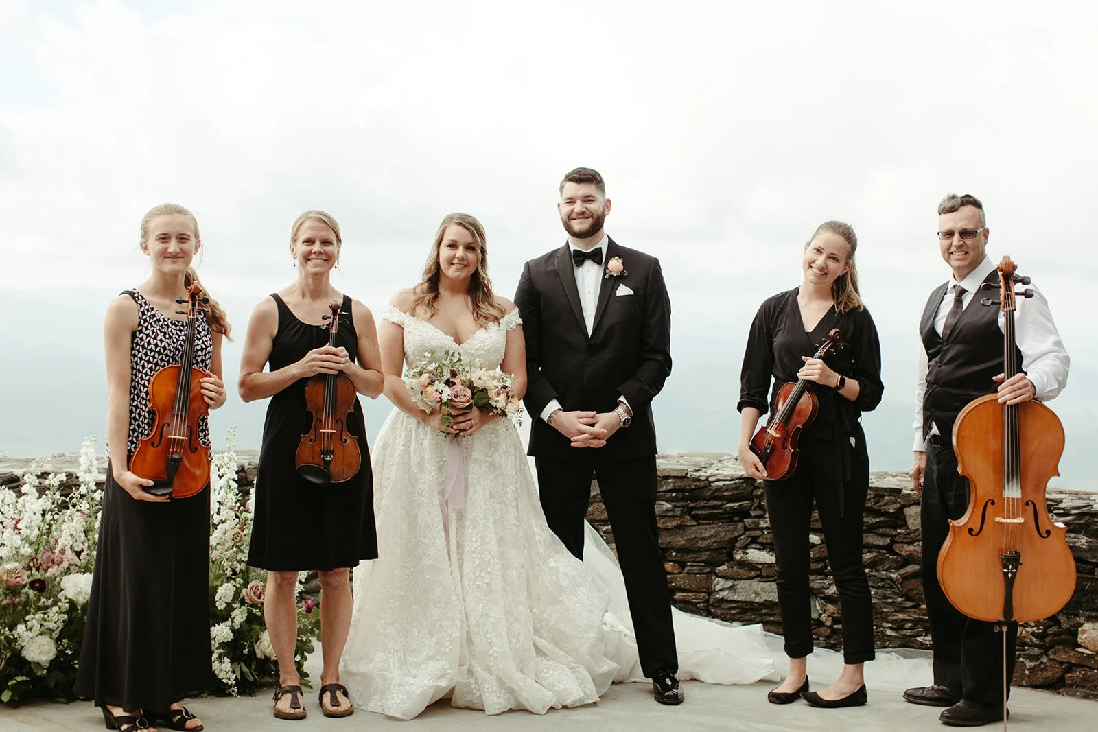 Bride and groom posing with ceremony musicians holding string instruments at a mountain overlook.
