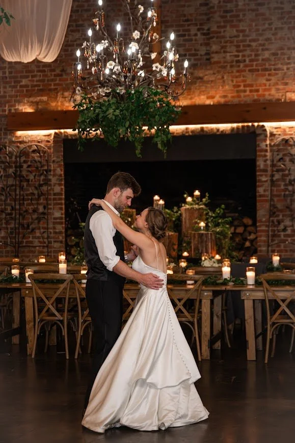 Bride and groom slow dance under a chandelier in a candlelit brick reception space.