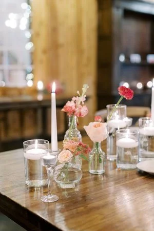 A softly lit wooden table features a romantic centerpiece with various small vases holding pink and coral flowers, surrounded by lit candles in glass holders and tall white tapers.