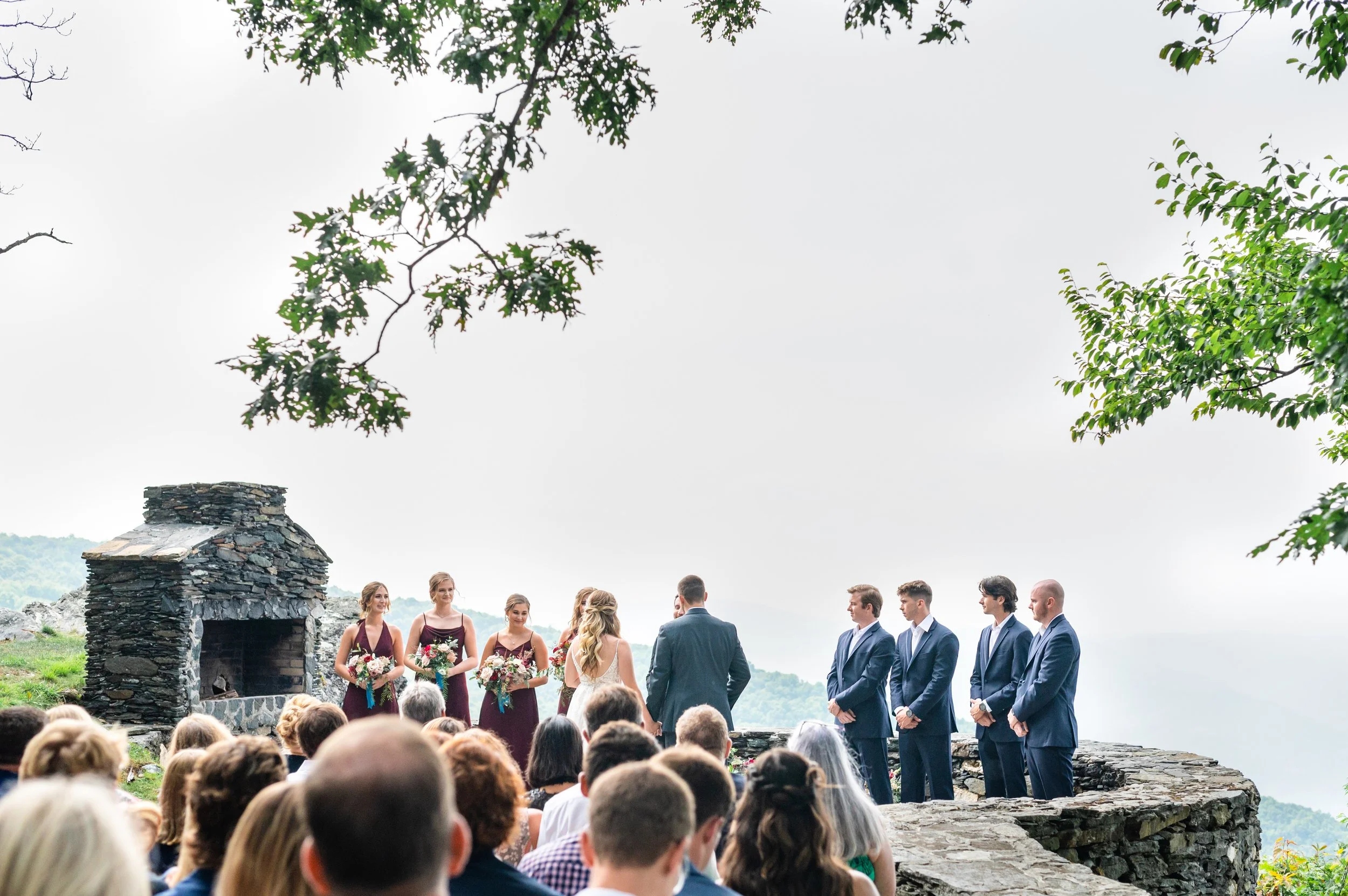 Bride and groom standing with their wedding party during an outdoor mountain overlook ceremony beside a stone fireplace.