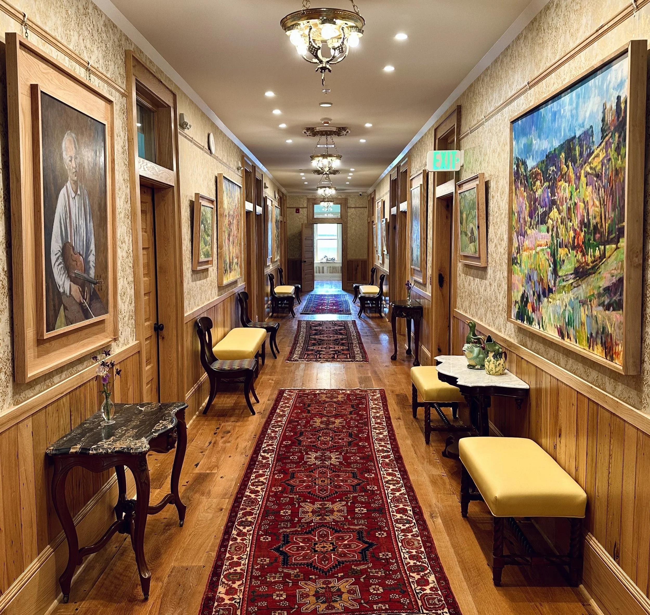 Warmly lit hotel hallway with framed artwork, wooden trim, benches, and patterned runner rugs.