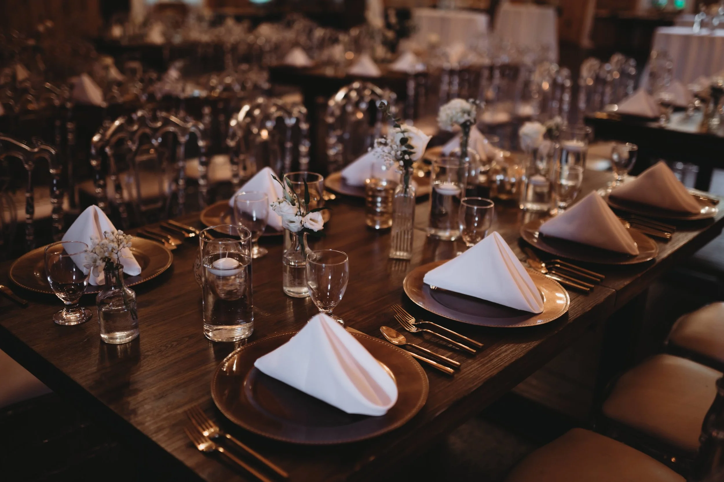 Elegant wedding reception table with folded white napkins, gold-rimmed plates, glassware, and candlelit floral bud vases