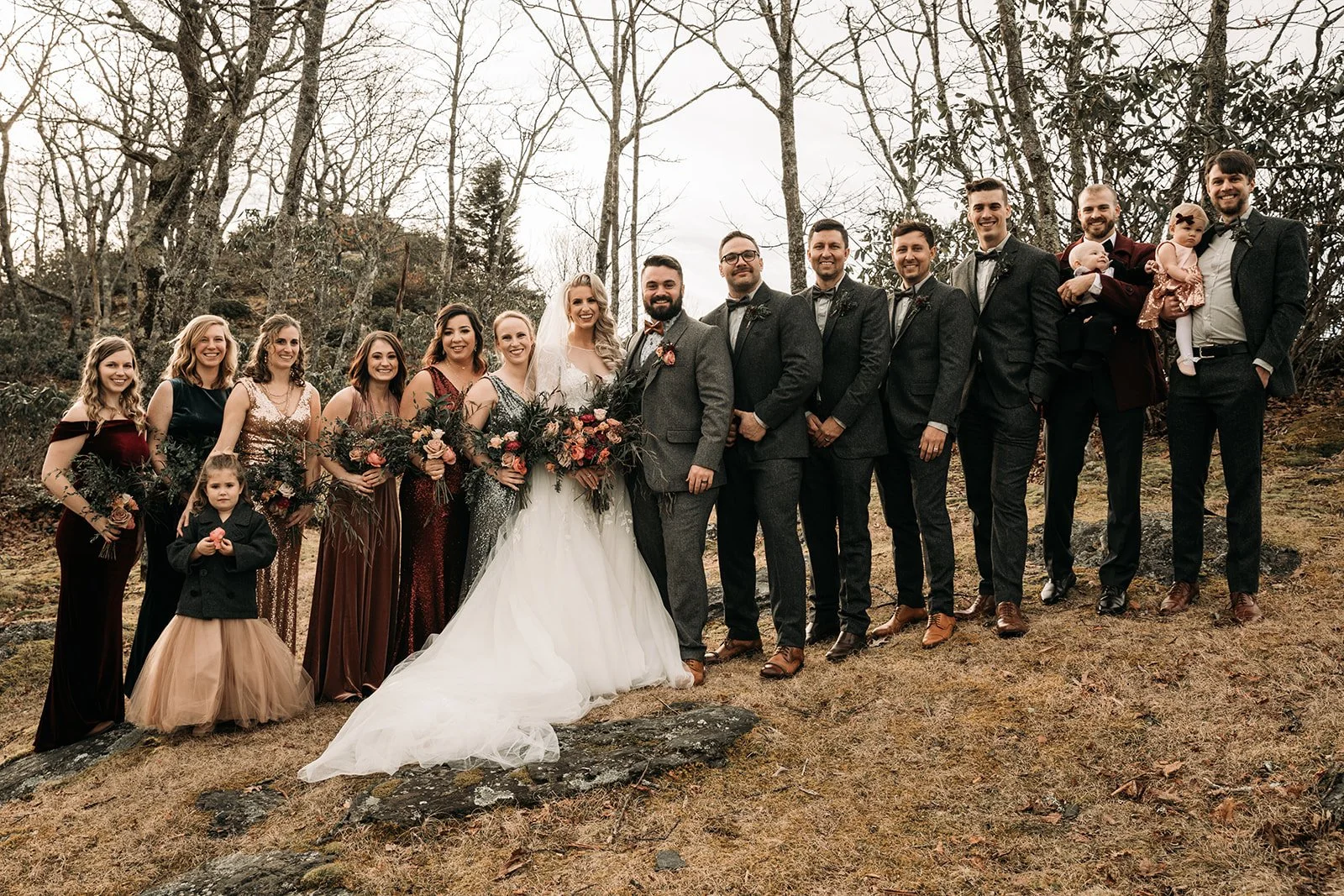 Wedding party posing outdoors in the woods with bridesmaids in warm tones and groomsmen in gray suits.