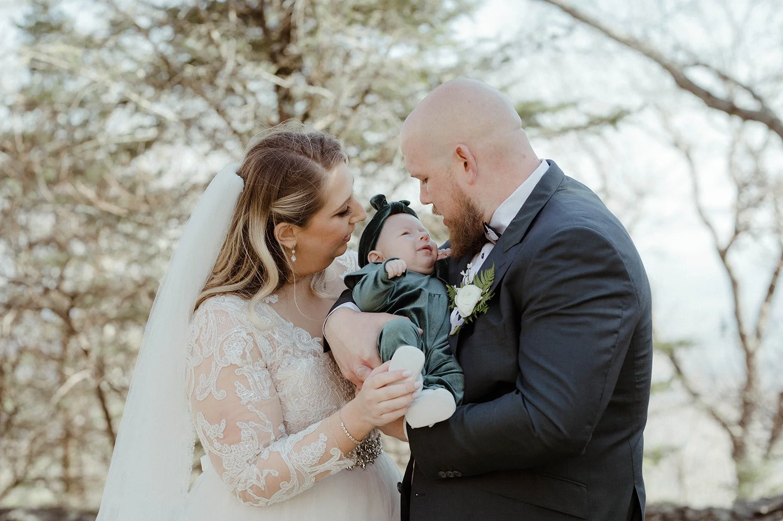 Bride and groom holding their baby during an outdoor family wedding portrait in soft natural light.