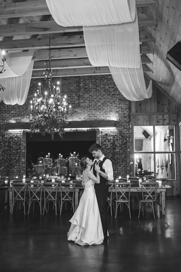 Black-and-white photo of bride and groom slow dancing in a rustic barn reception under chandeliers and ceiling draping