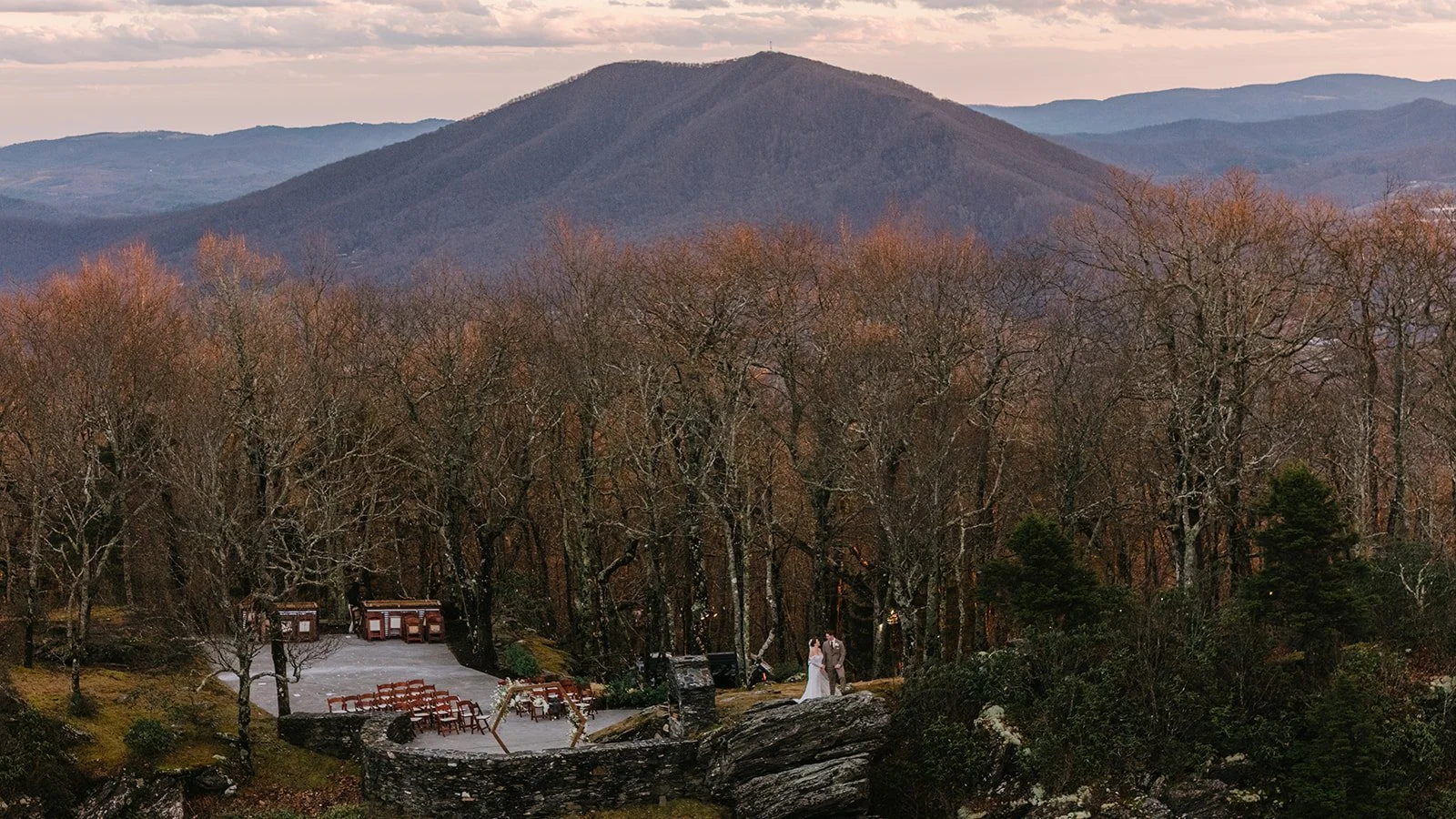 Wide view of a mountain wedding ceremony site with the bride and groom standing near rocky cliffs at sunset.