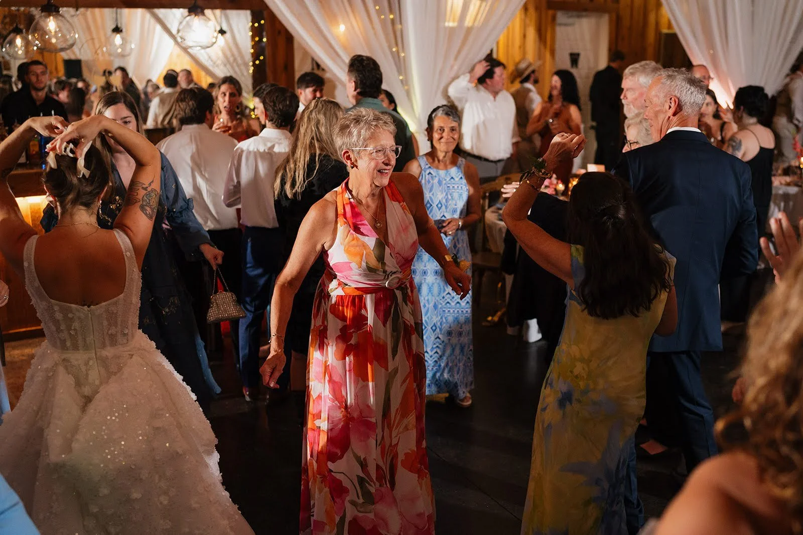 Wedding guests dancing during a lively rustic reception with white draping and warm indoor lighting.