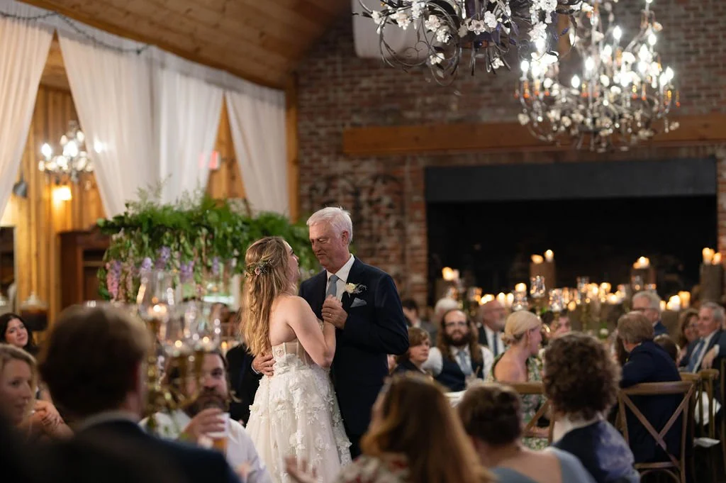 Bride dancing with her father beneath a chandelier during a warm candlelit wedding reception.