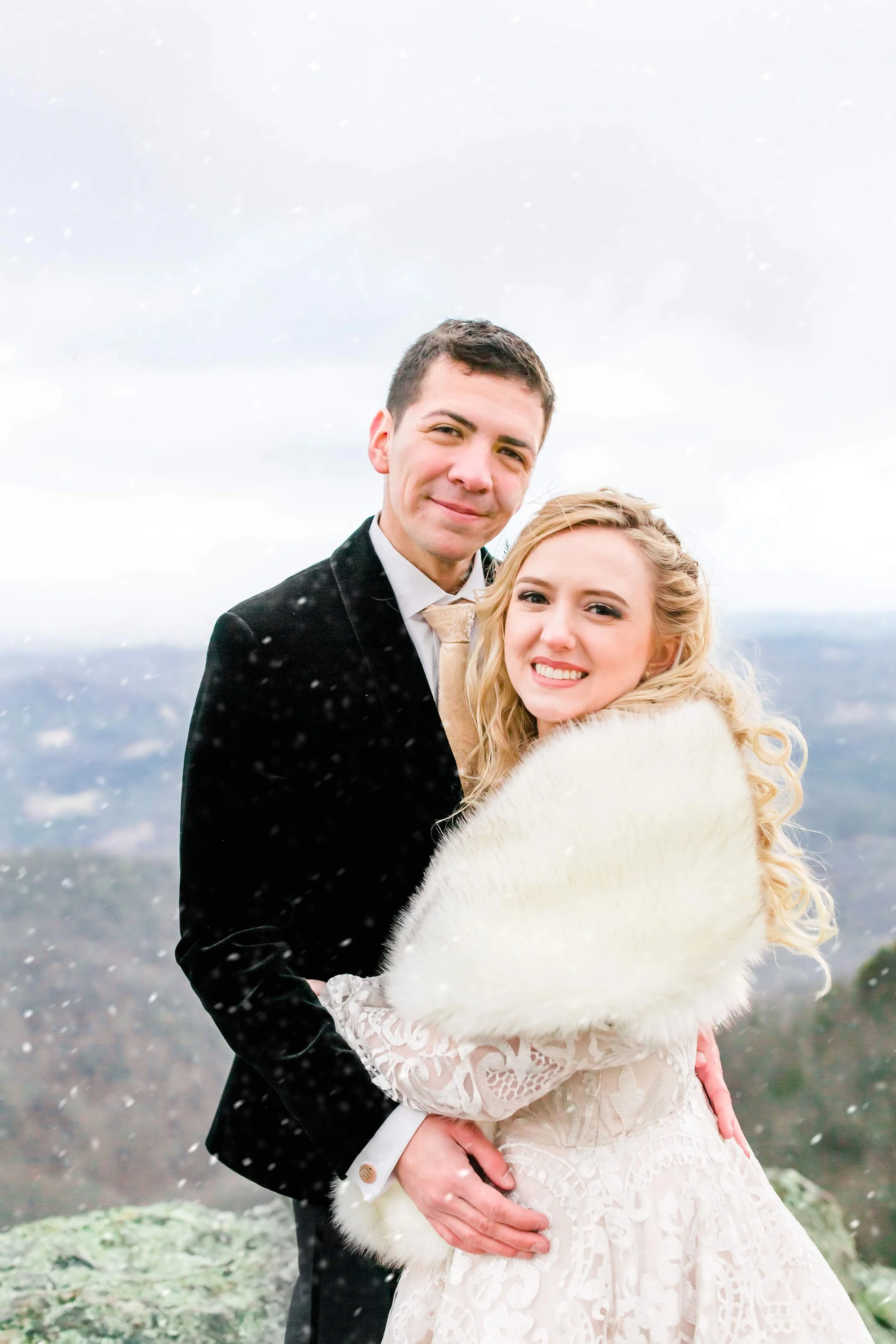 Winter wedding couple smiling in light snowfall with mountain views, bride wrapped in a white fur shawl