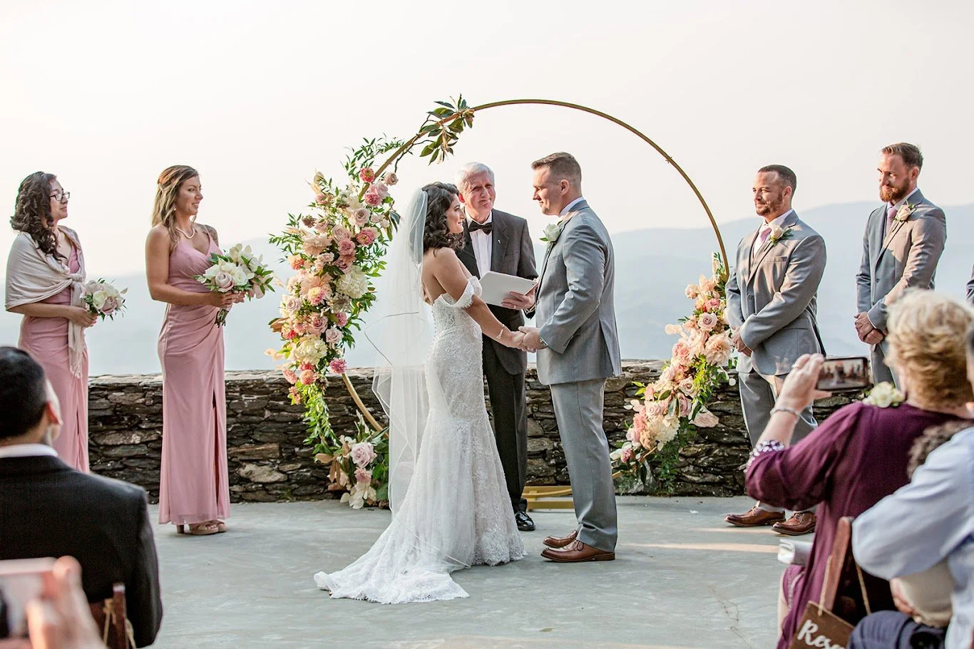 Bride and groom holding hands during vows under a circular floral arch with mountain views