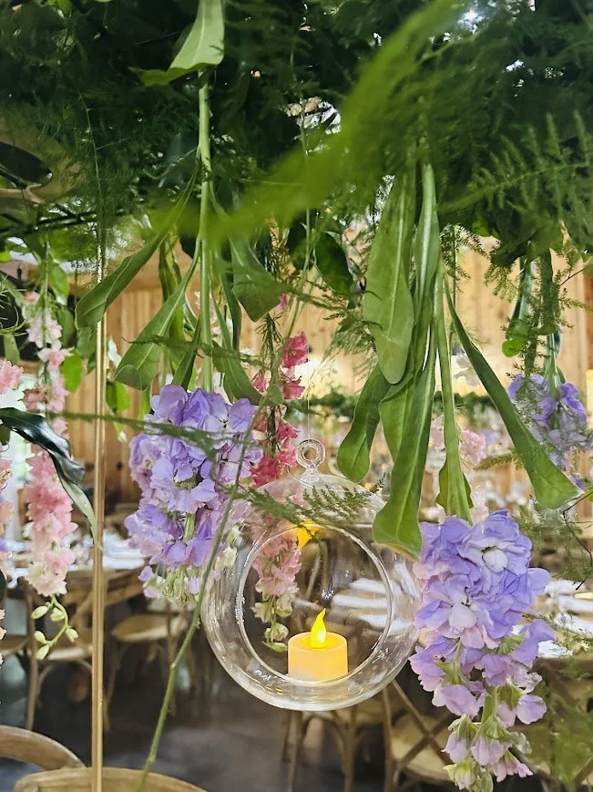 Hanging glass orb with a candle beneath lush greenery and purple hydrangea for an outdoor reception decor detail.