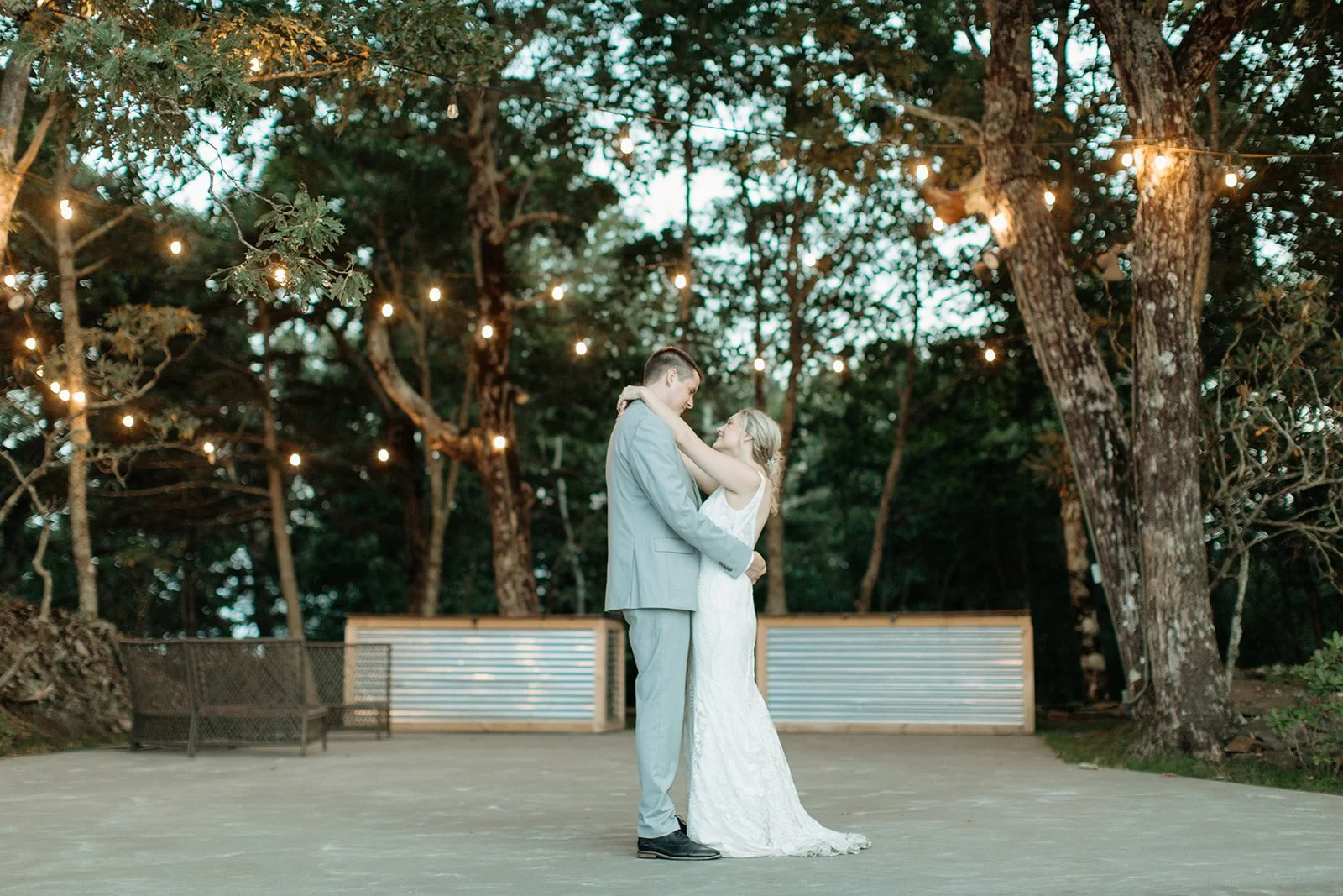 Bride and groom sharing their first dance outdoors beneath glowing string lights and trees at dusk.