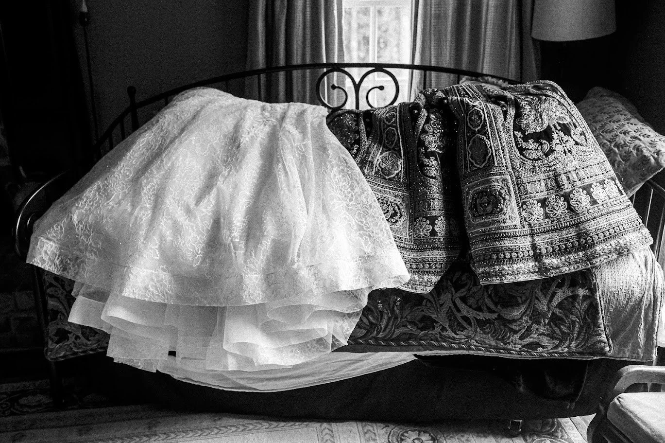 Black-and-white photo of a wedding dress and an embroidered outfit draped over a bed near a window.