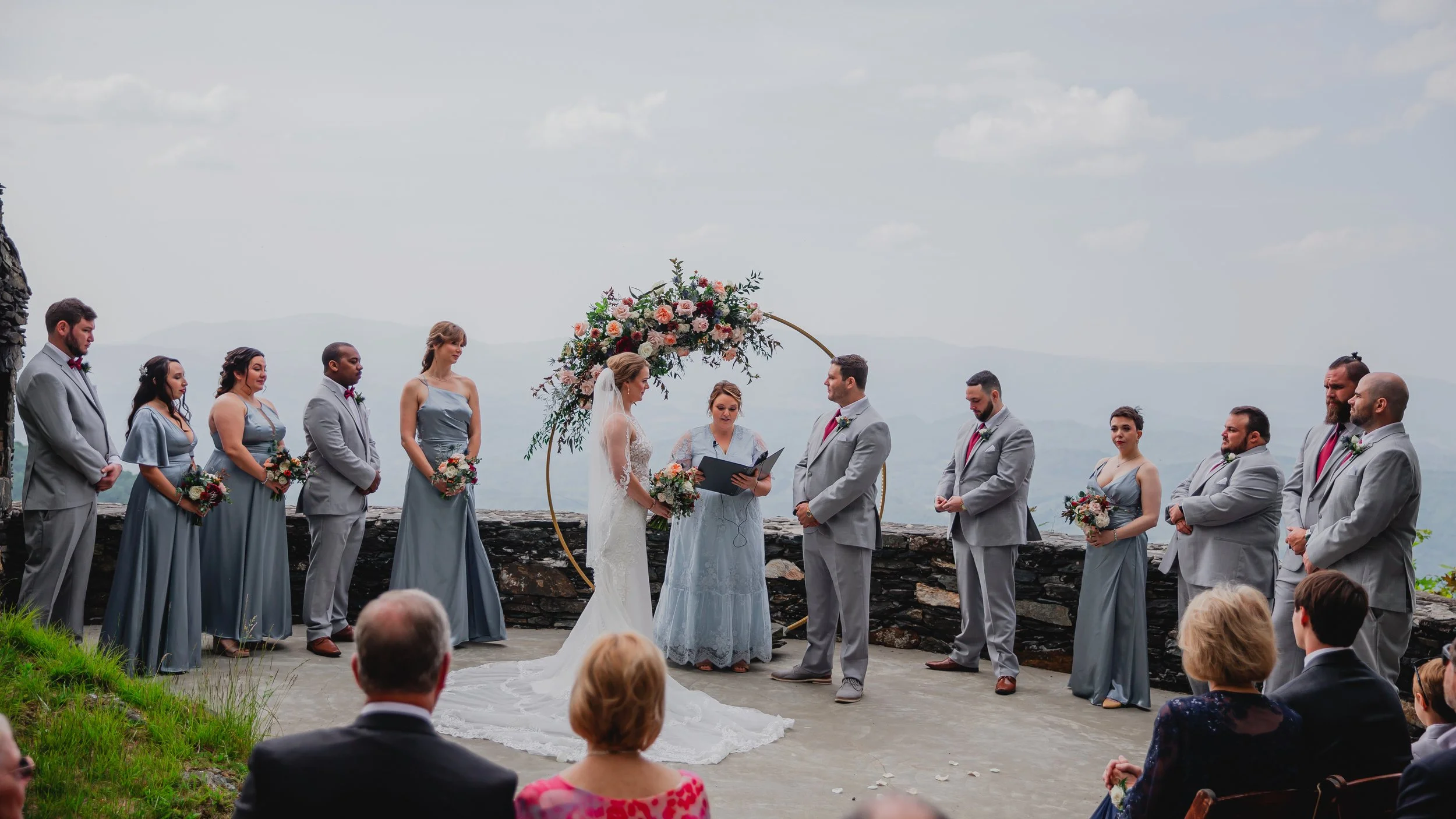 Outdoor ceremony with a floral arch and wedding party in grey attire as the couple stands at the altar with mountain views.
