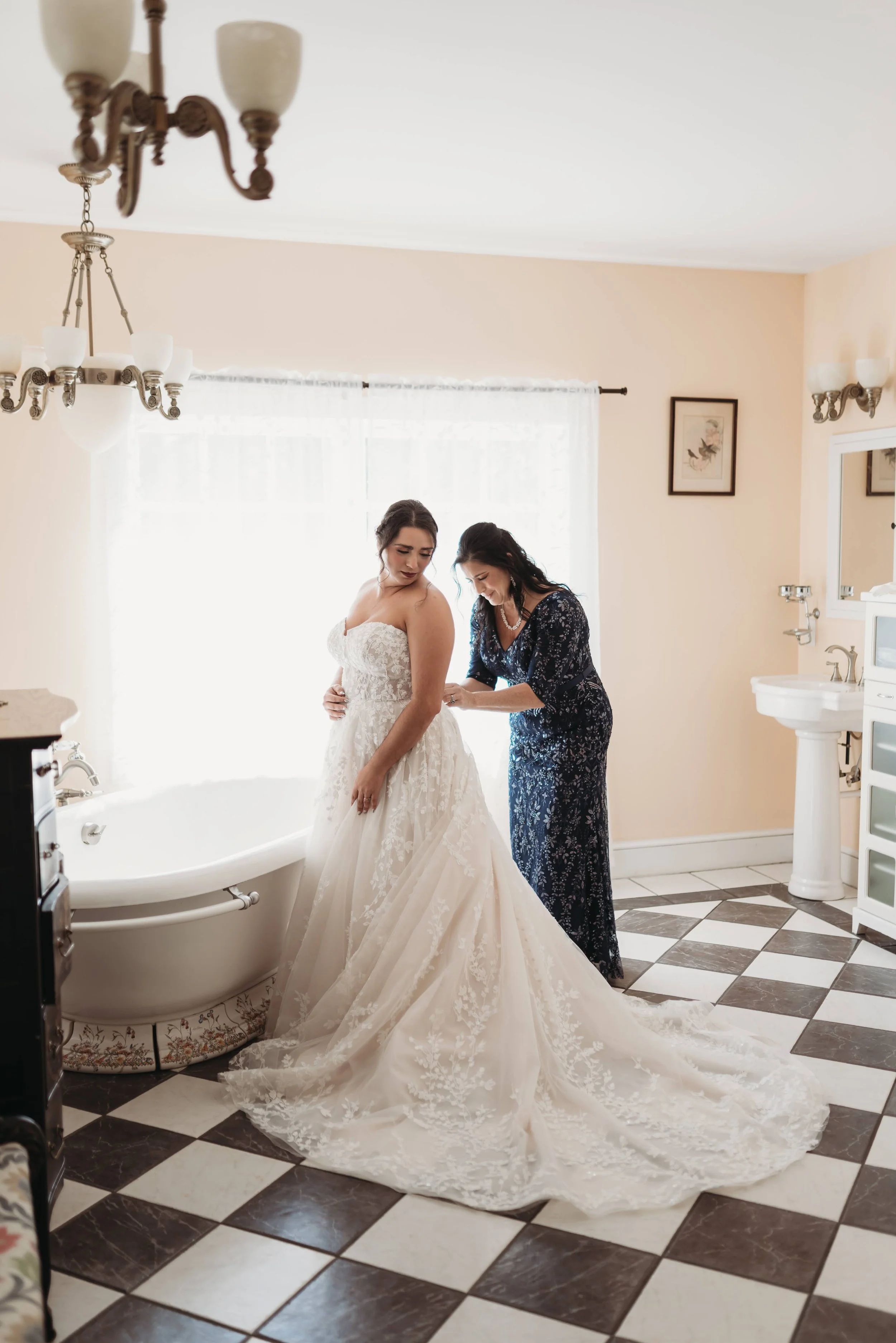 Bride in a lace wedding gown as a helper makes final adjustments in a bright bathroom.