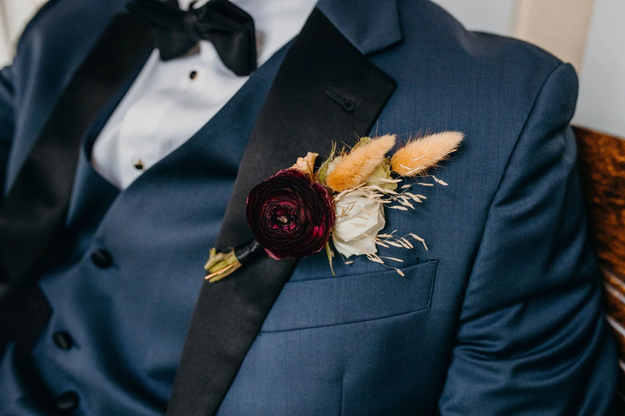 Closeup of a groom’s boutonniere with a burgundy flower and dried accents on a blue suit.