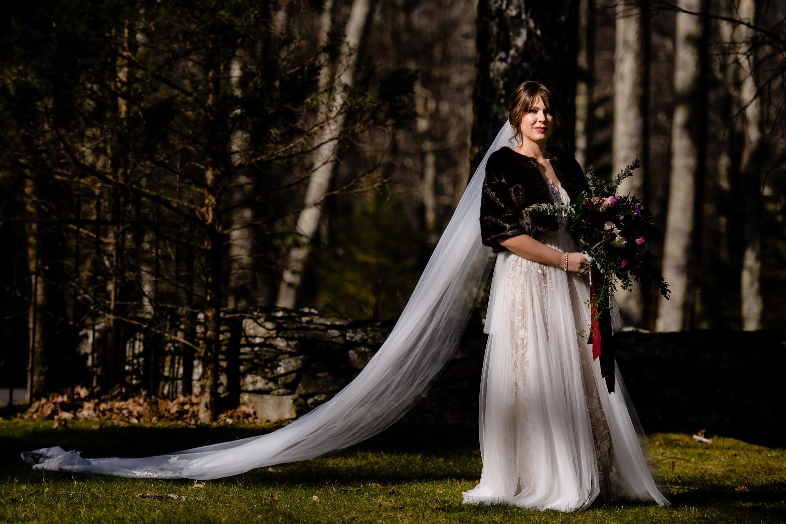 Bride wearing a fur wrap with a long veil, holding a bouquet outdoors in a wooded setting.