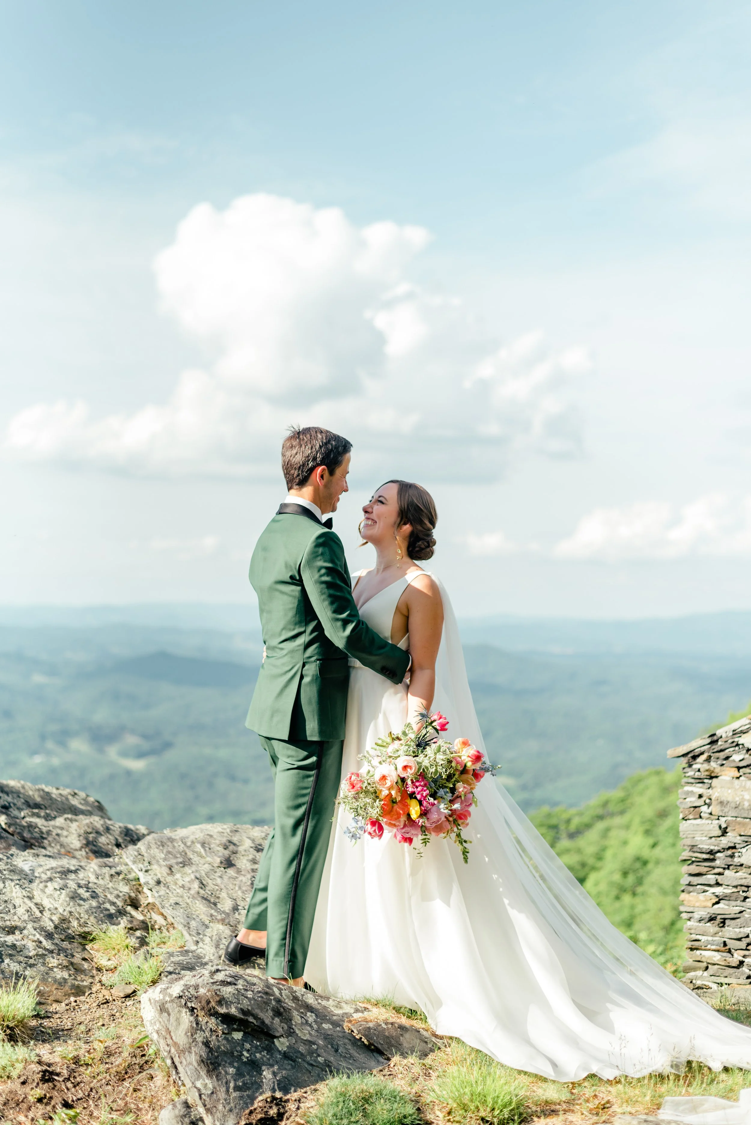 Bride and groom standing together on a mountain overlook with the groom in a green suit and the bride holding a colorful bouquet.