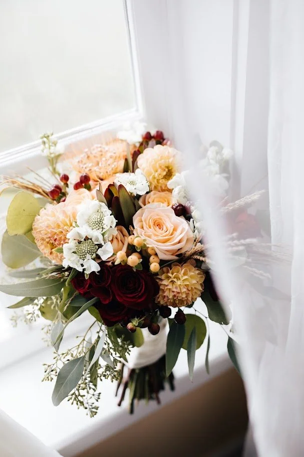 Closeup of a fall wedding bouquet with roses, dahlias, and burgundy blooms near a bright window.