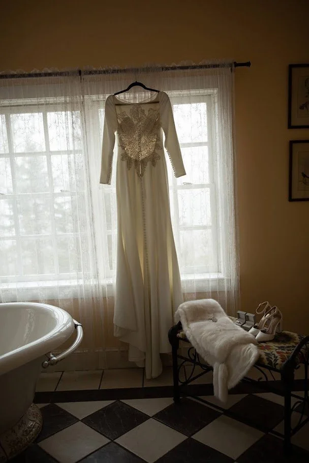 Wedding dress hanging in front of a bright bathroom window with a vintage tub nearby.