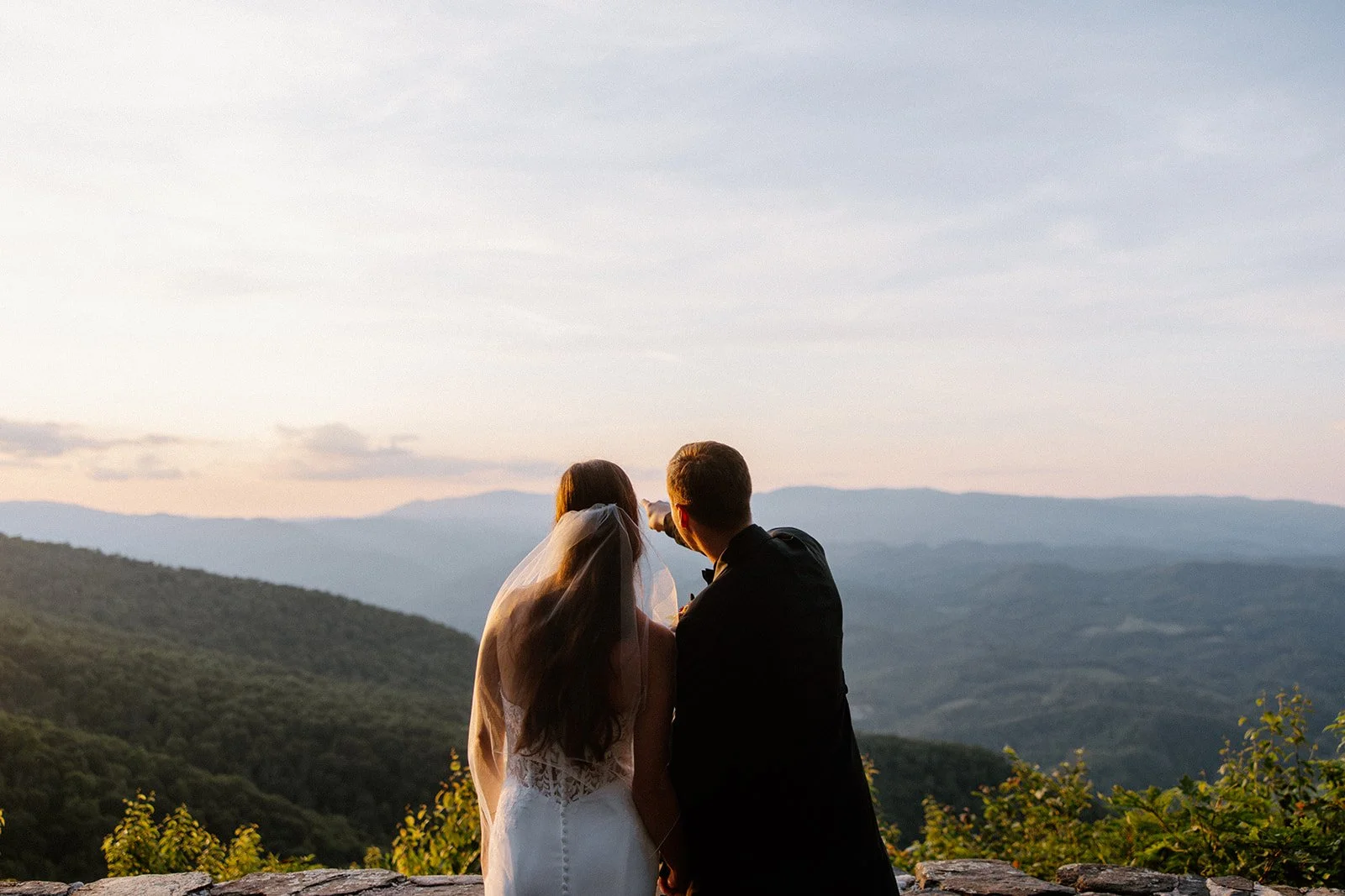 Bride and groom viewed from behind at sunset looking out over layered mountain ridges.