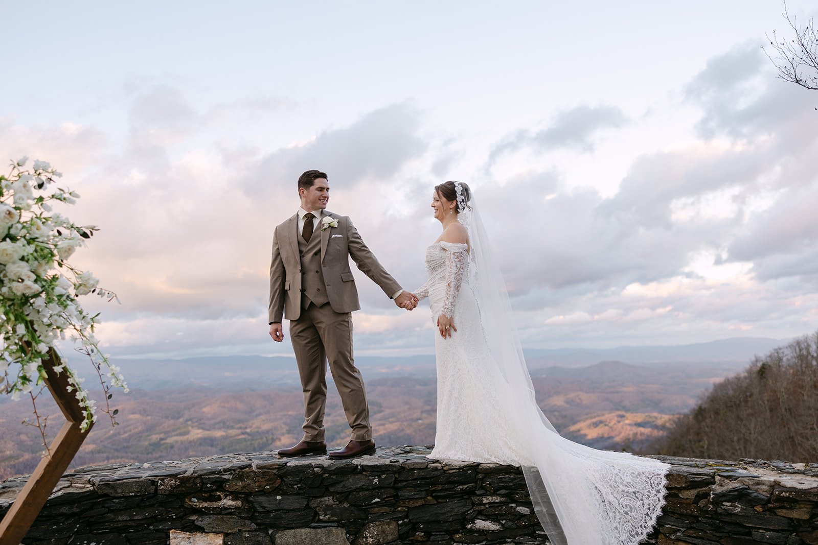 Bride and groom holding hands on a stone wall at a mountain overlook with dramatic clouds and valley views.