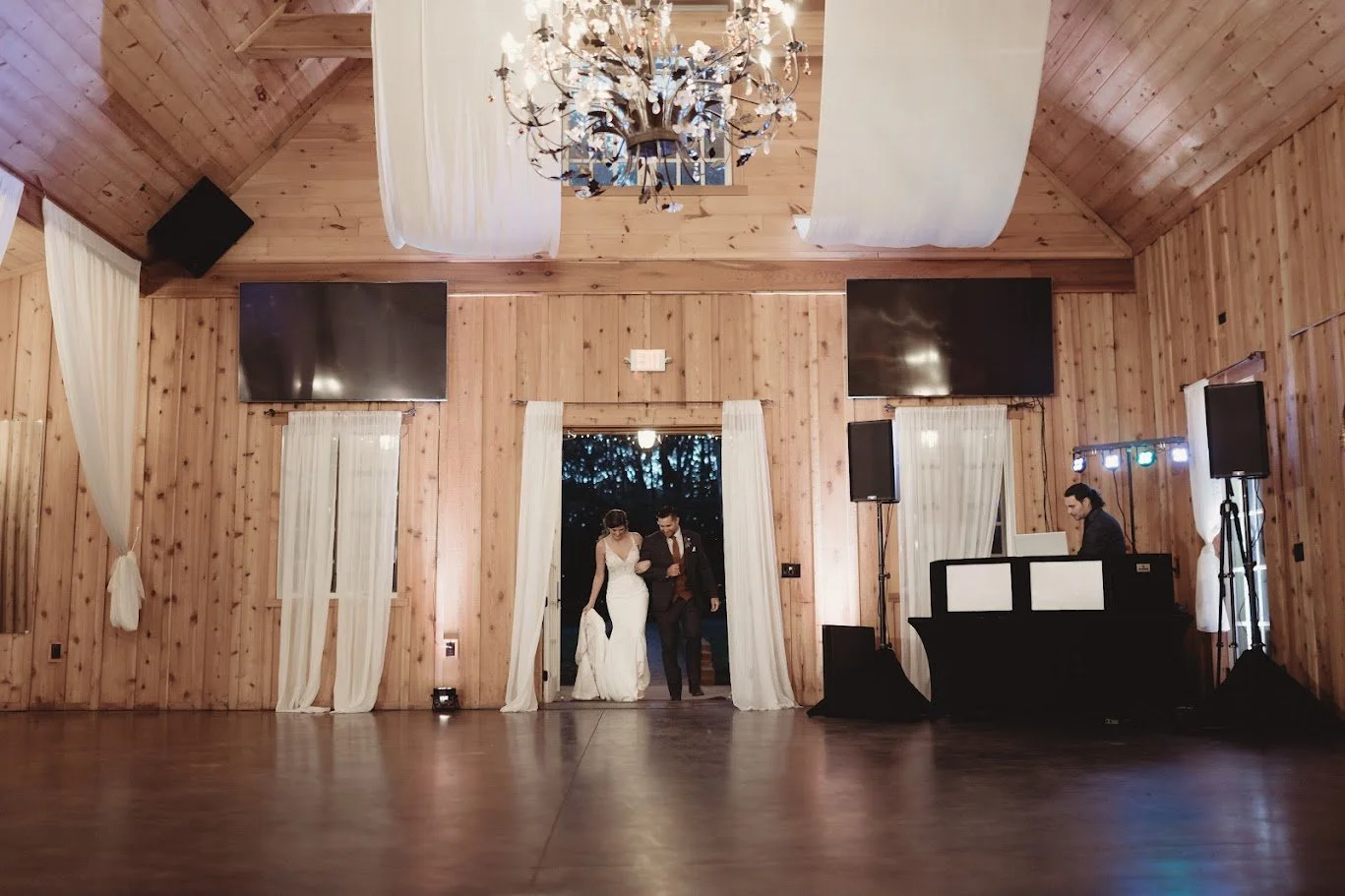 Bride and groom entering a barn reception through white-draped double doors under a chandelier