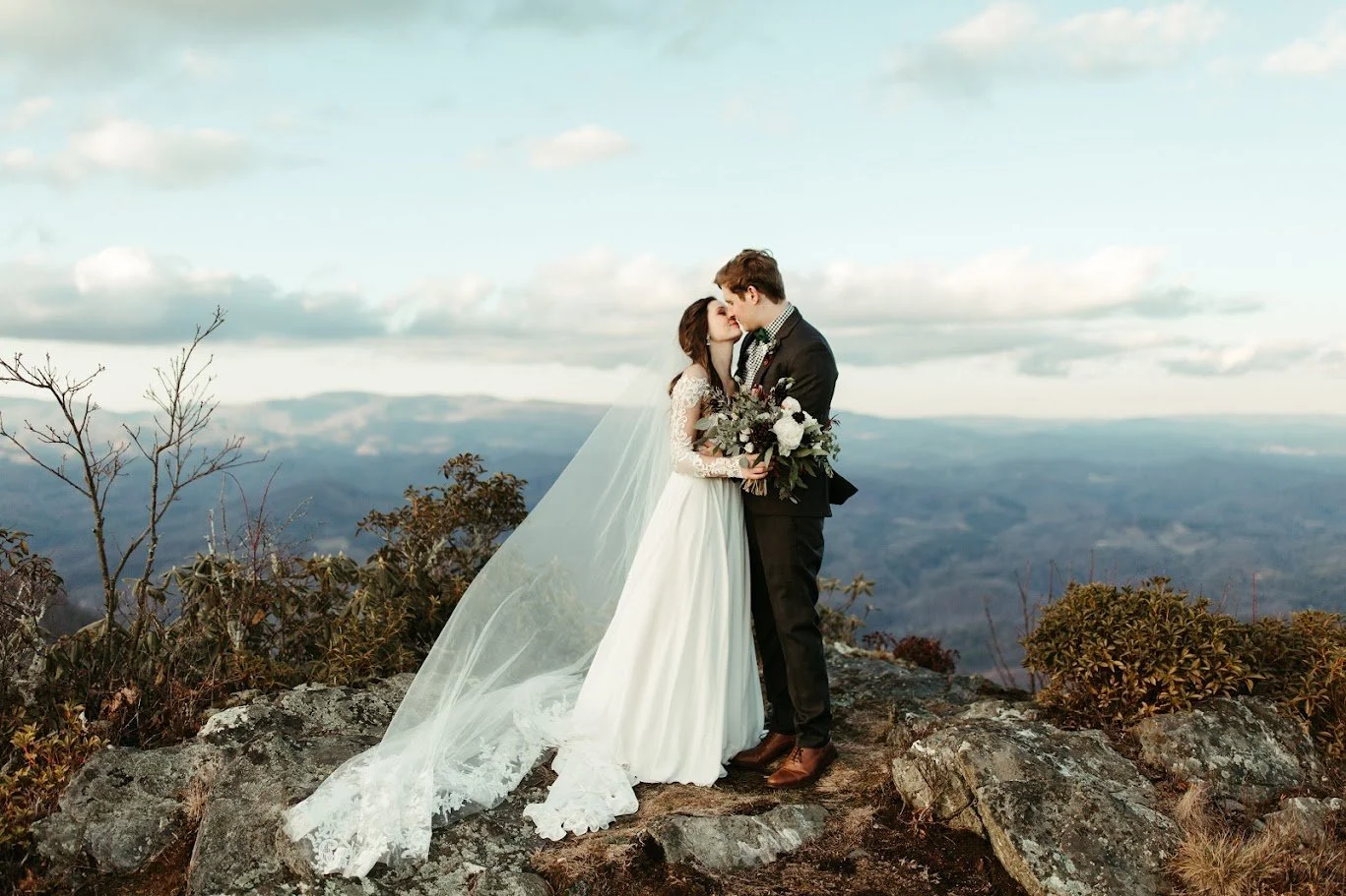 Bride and groom kissing on a rocky mountain overlook with a long veil and scenic blue ridge views.