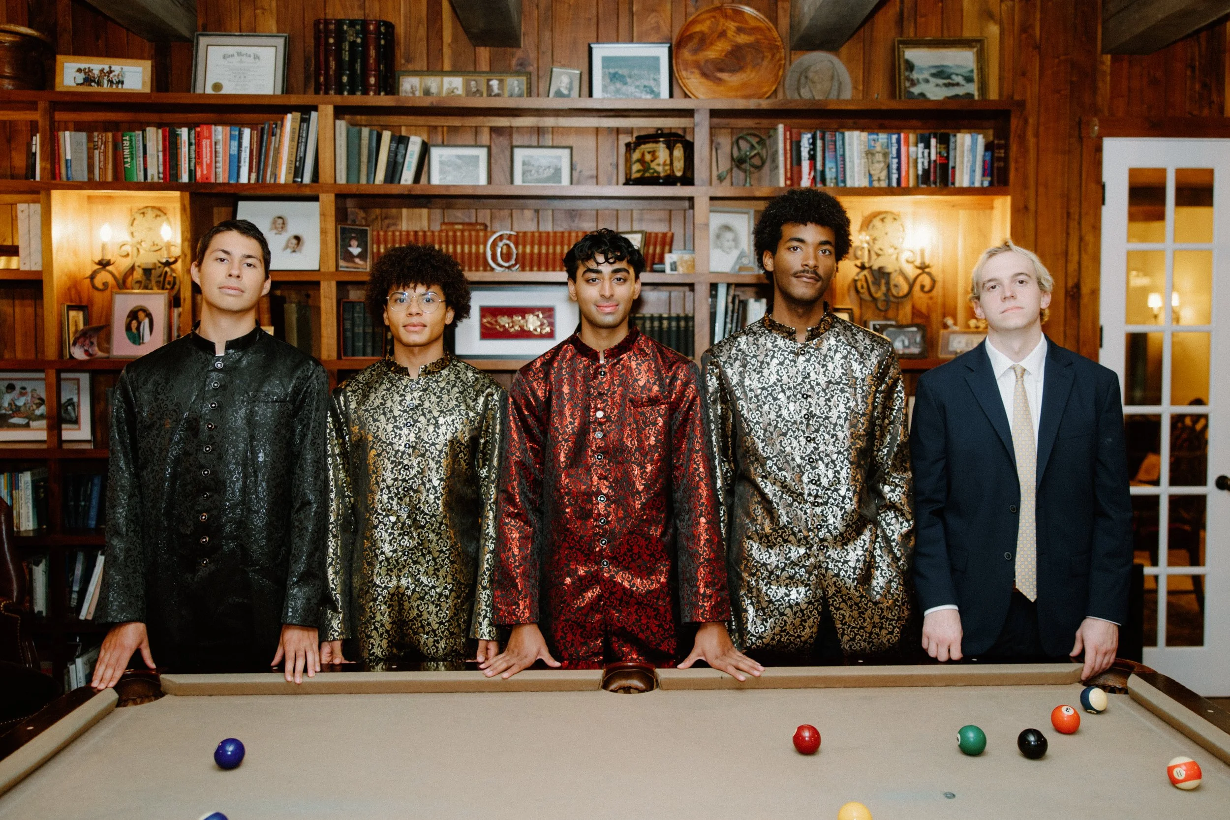 Five men pose behind a pool table in a wood-paneled library room, wearing patterned sherwanis and a suit.
