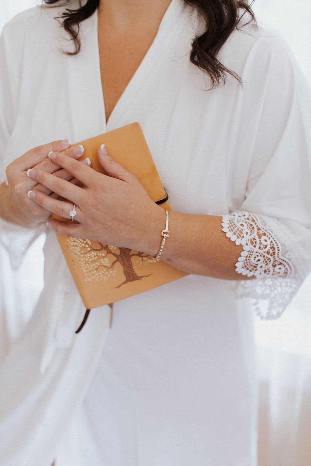 Closeup of the bride in a white lace-trim robe holding a vow journal, showing her engagement ring and bracelet.