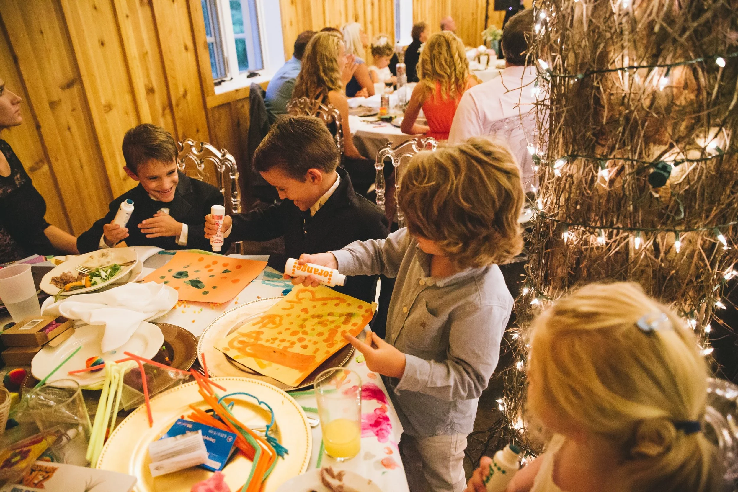 Children painting and playing at a kids activity table during a rustic wedding reception with string lights and dinner guests nearby.