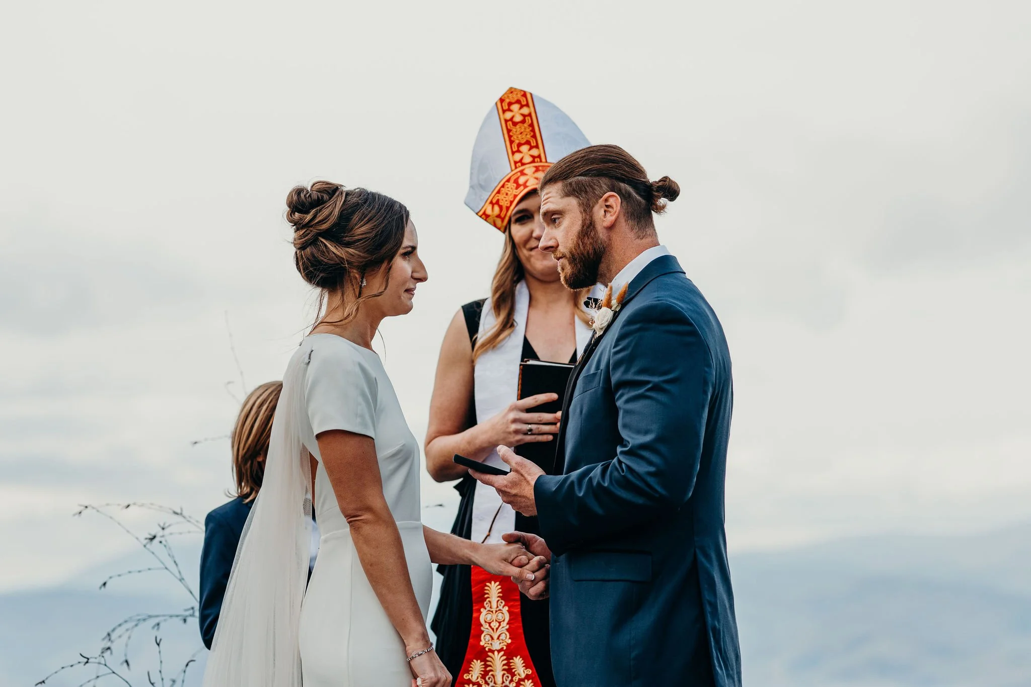 Couple exchanging vows during an outdoor mountain ceremony, groom in a blue suit and officiant behind them.